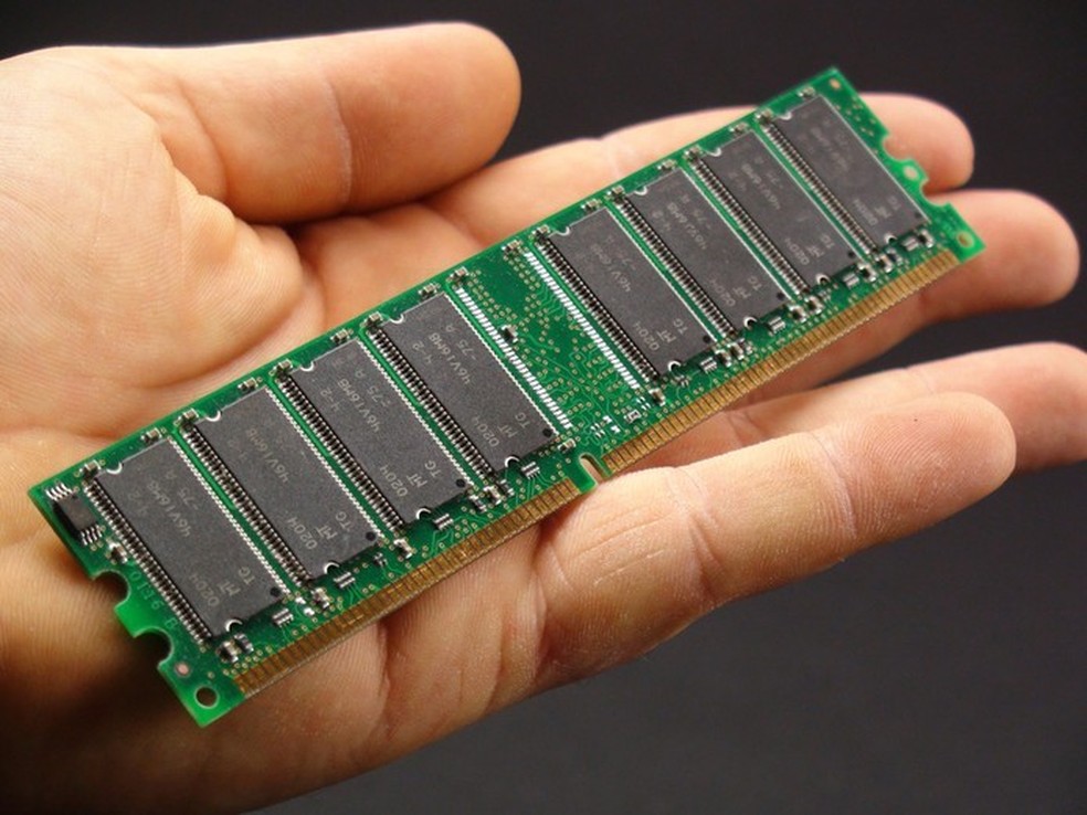  A imagem mostra uma mão segurando um módulo de memória RAM. O módulo é uma placa retangular verde com vários chips pretos alinhados em duas fileiras. Na parte inferior, há uma série de contatos dourados que se conectam à placa-mãe do computador. O fundo da imagem é preto, destacando o módulo de memória na mão.