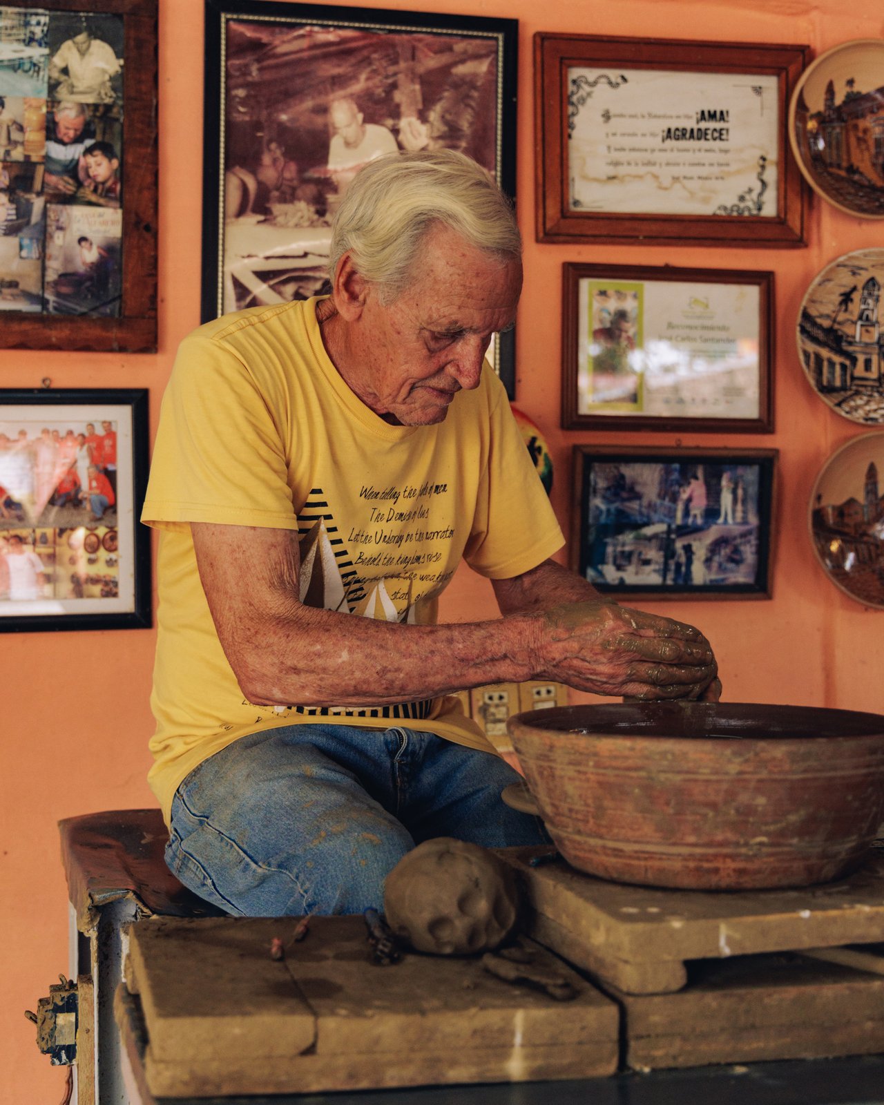 An older potter shaping clay at his wheel, framed photos on the wall, Trinidad, Cuba