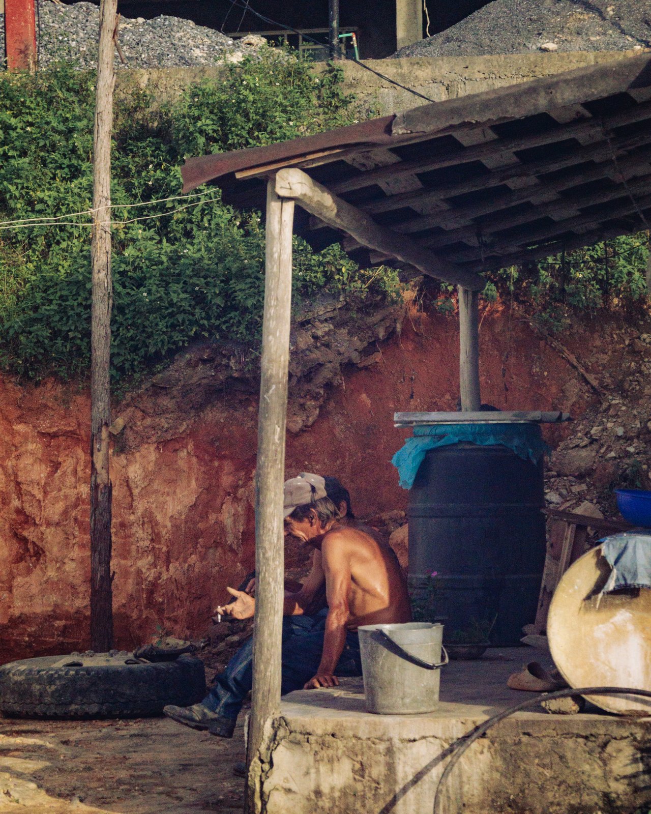 A shirtless man resting by an oil drum against a red earthen wall, rural Cuba