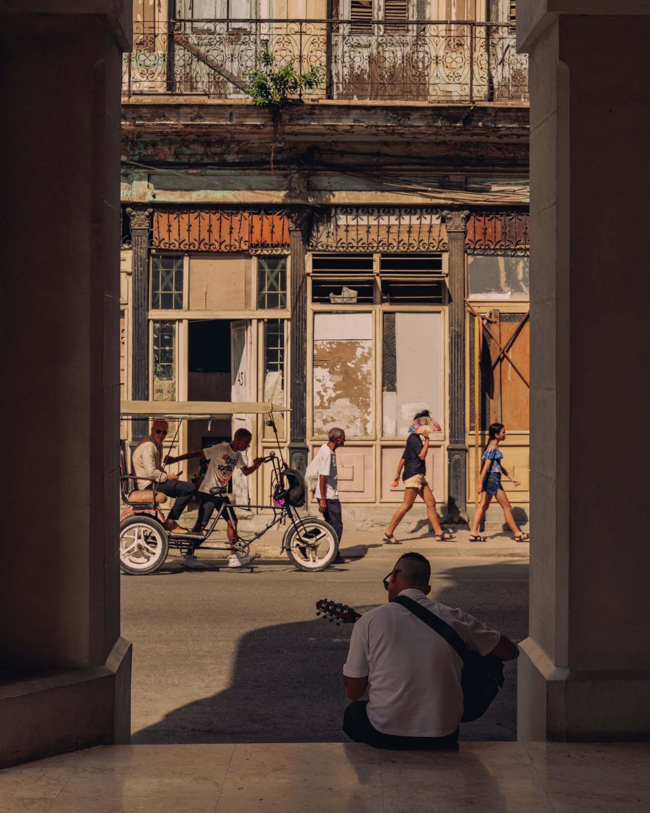 Bici-taxis and pedestrians under the arcade of a Havana street, a man with a guitar seated, Cuba