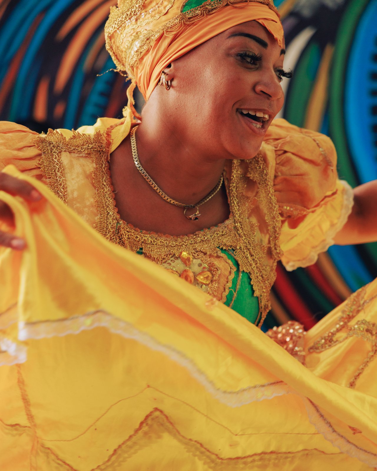 A dancer in a yellow dress and headwrap twirling her skirt before a colourful mural, Cuba