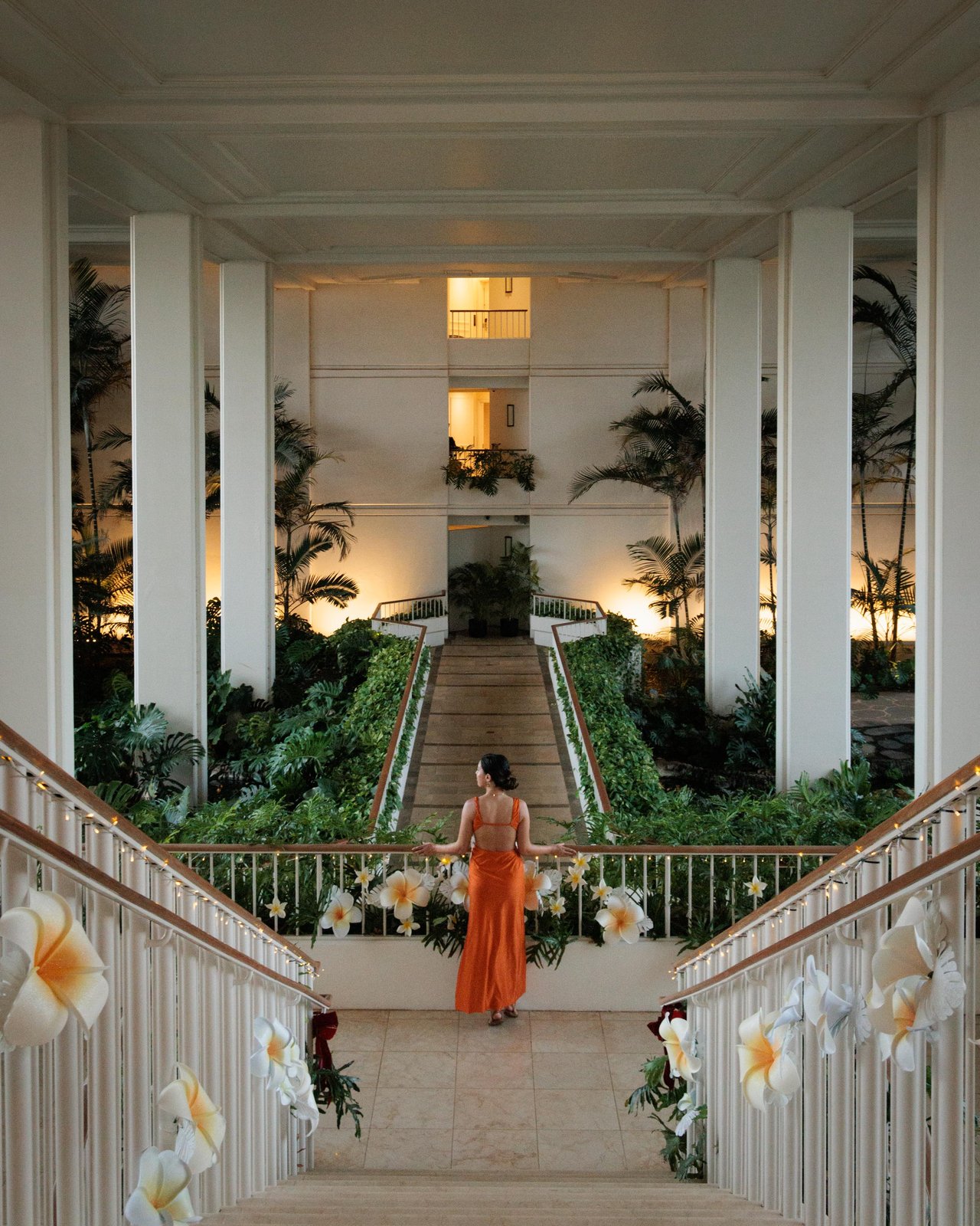 A woman in an orange dress on a grand staircase draped with plumeria garlands at dusk, Four Seasons Oʻahu