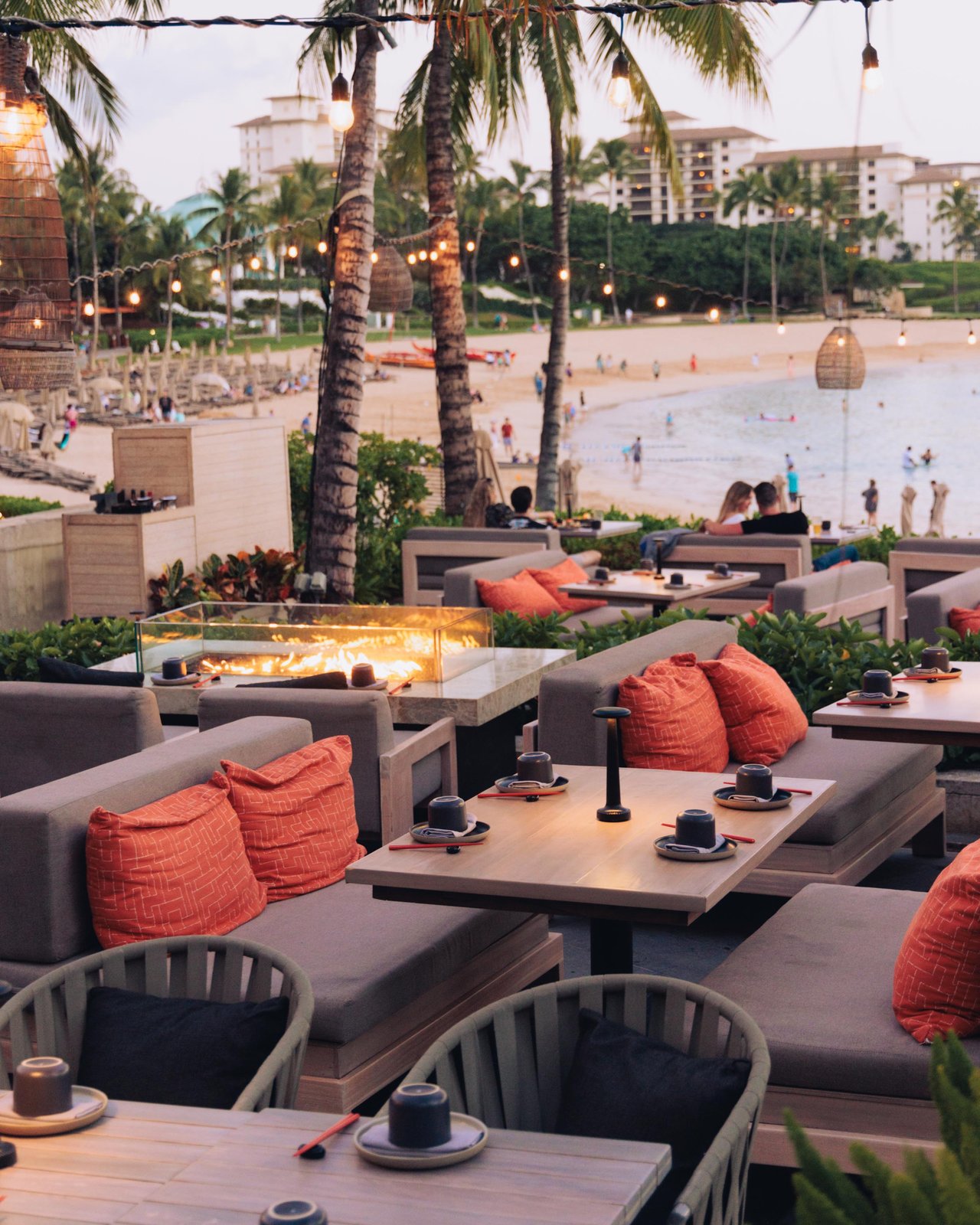 A beachfront terrace lounge with fire tables and orange cushions, palms and the lagoon beyond, Ko Olina