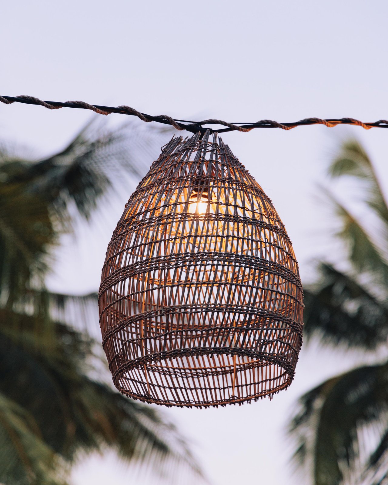 A woven rattan pendant lamp glowing among palm fronds at dusk, Four Seasons Oʻahu