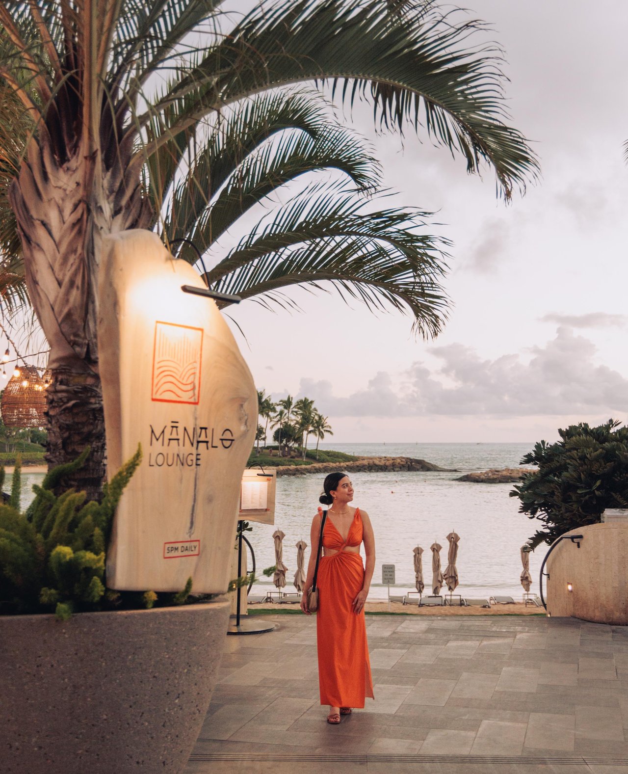 A woman in an orange dress by the Mānalo Lounge sign, a palm and the ocean cove behind, Ko Olina