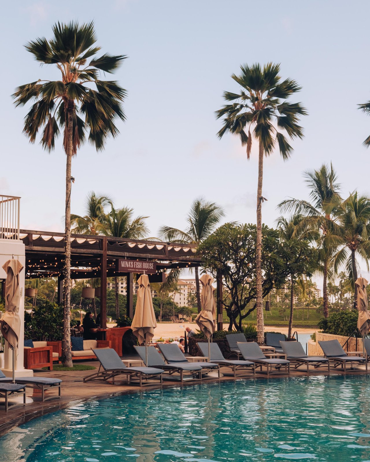 The resort pool at dusk with chaise lounges and palms, Mina's Fish House behind, Ko Olina