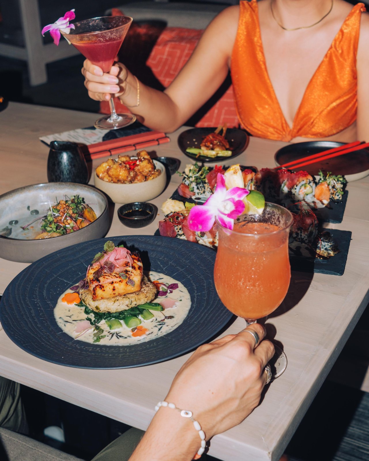 A woman in an orange dress raising a cocktail over a sushi dinner, Four Seasons Oʻahu