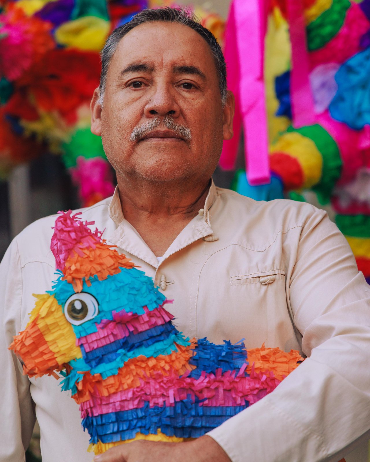 A man holding a bright handmade piñata before a wall of hanging piñatas, Mexico