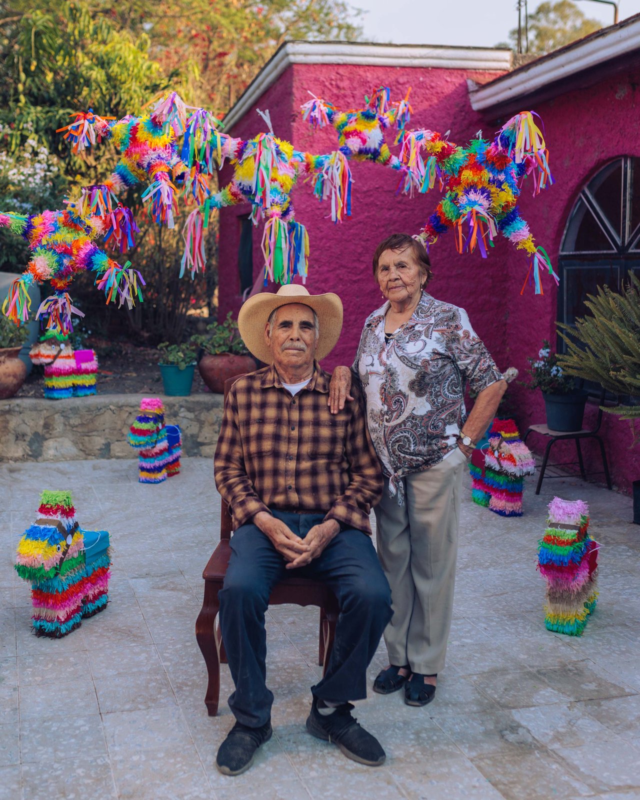 An older couple in a pink-walled courtyard hung with piñatas, the man seated in a hat, Mexico