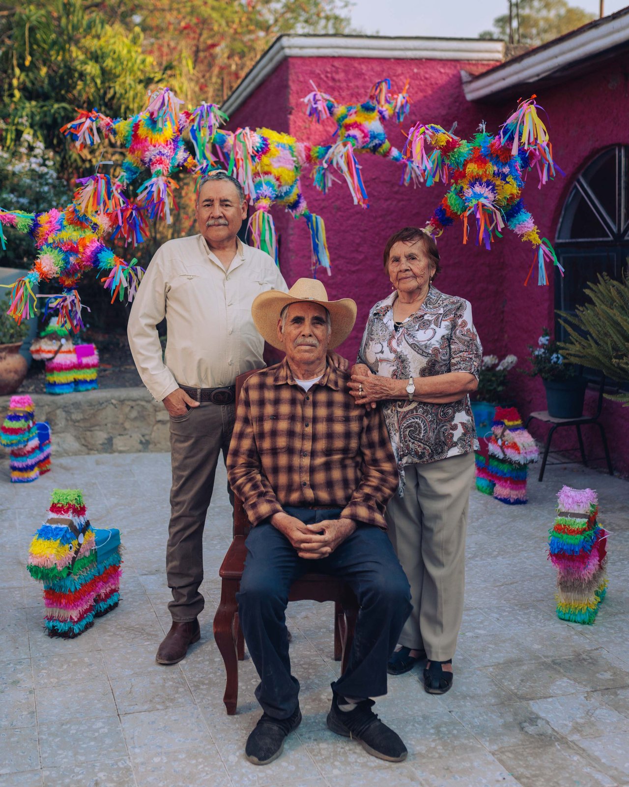 A family portrait in a pink courtyard of piñatas, a seated man in a straw hat, Mexico