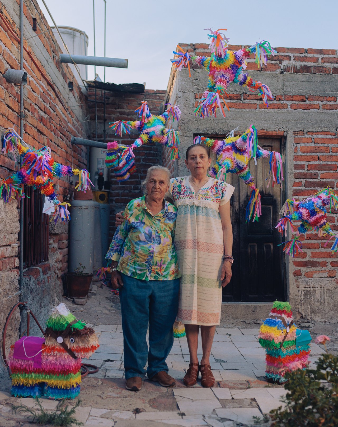 An older couple in a brick courtyard hung with piñatas, a donkey piñata beside them, Mexico
