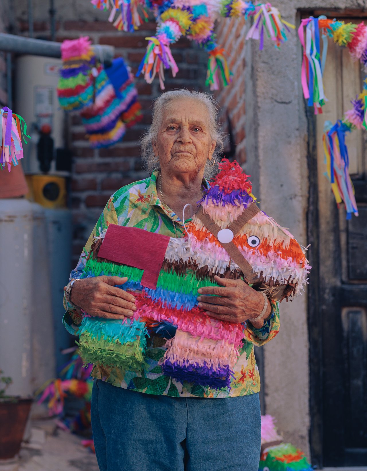 An older woman holding a donkey piñata in a brick courtyard of hanging piñatas, Mexico