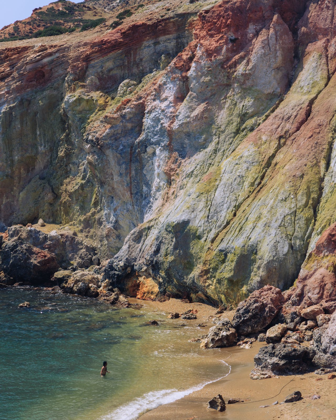 Colourful mineral cliffs above a turquoise cove with a swimmer, Milos, Greece