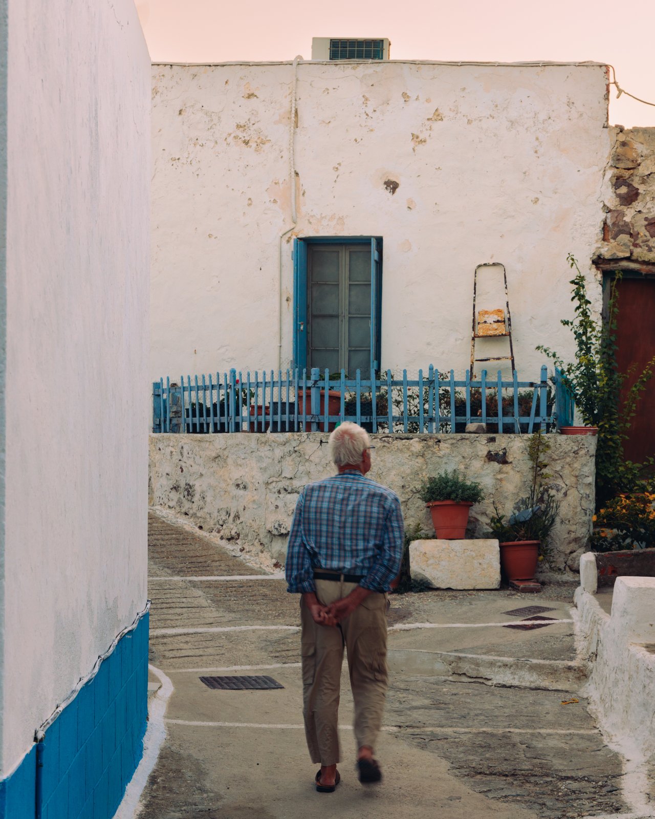 An older man walking a whitewashed village lane with blue railings and potted plants, Greece