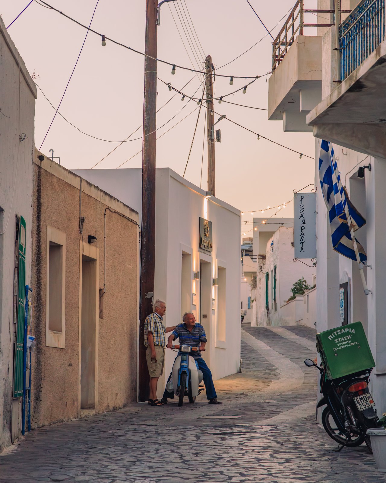 Two men chatting in a narrow whitewashed alley with a Greek flag at dusk, Greece