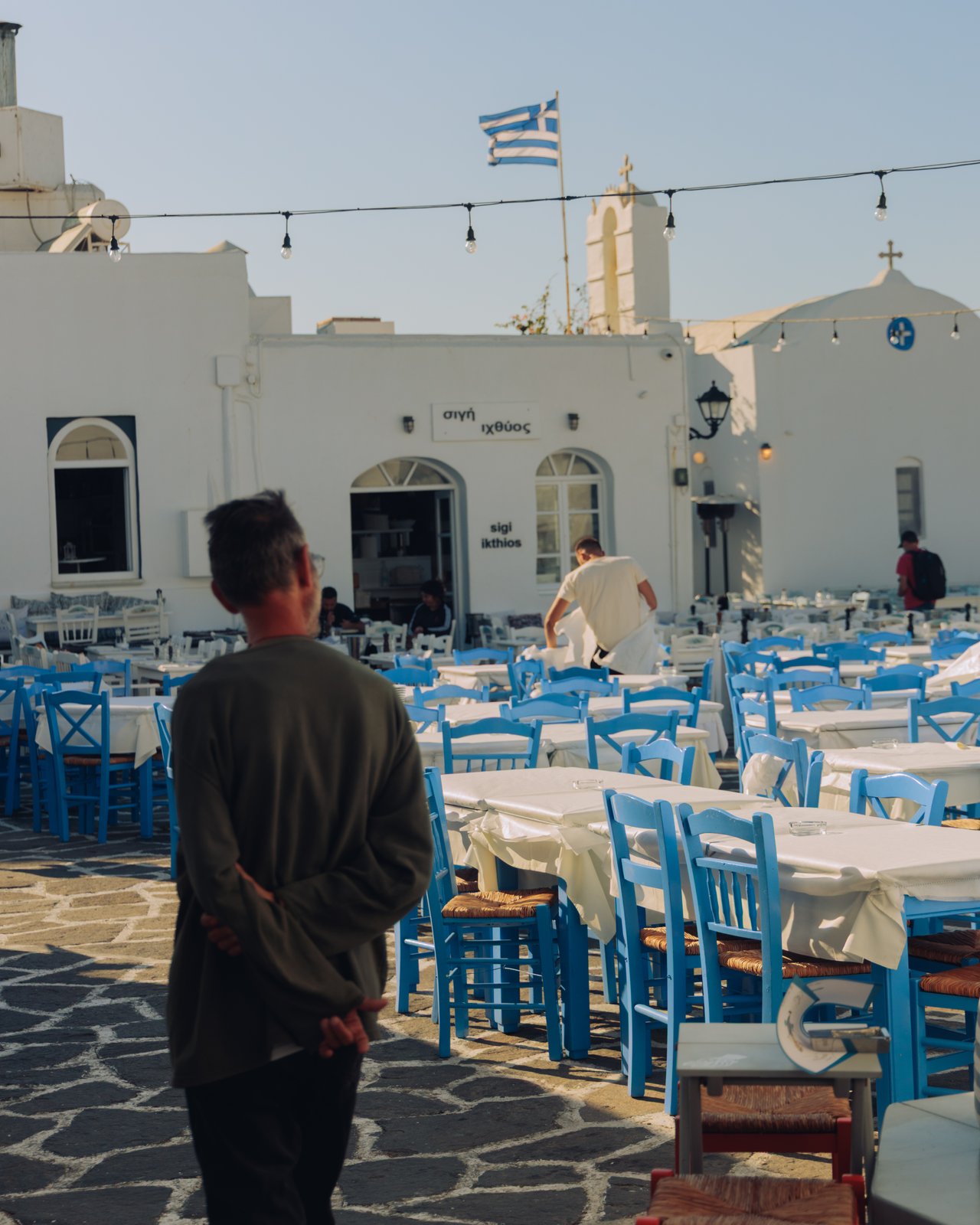 A man before rows of empty blue tavern chairs set out in a whitewashed square, Greece