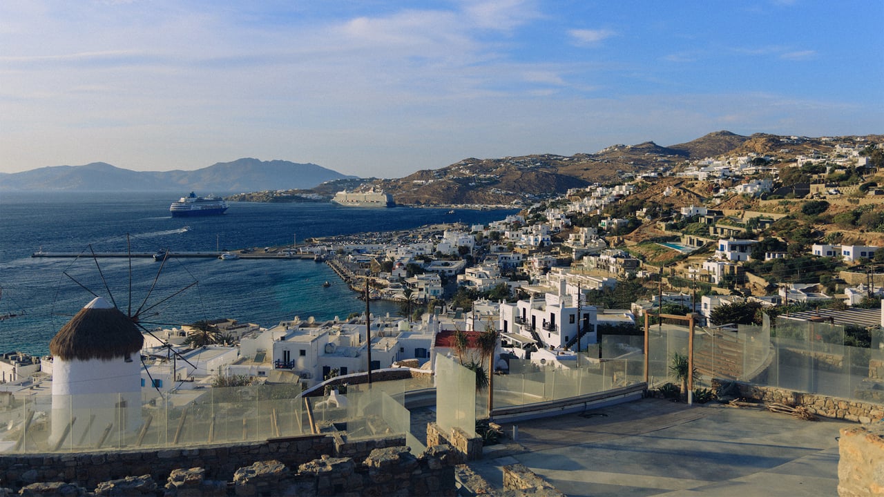 An elevated view over a Cycladic town and harbour at golden hour, a windmill in the foreground, Greece