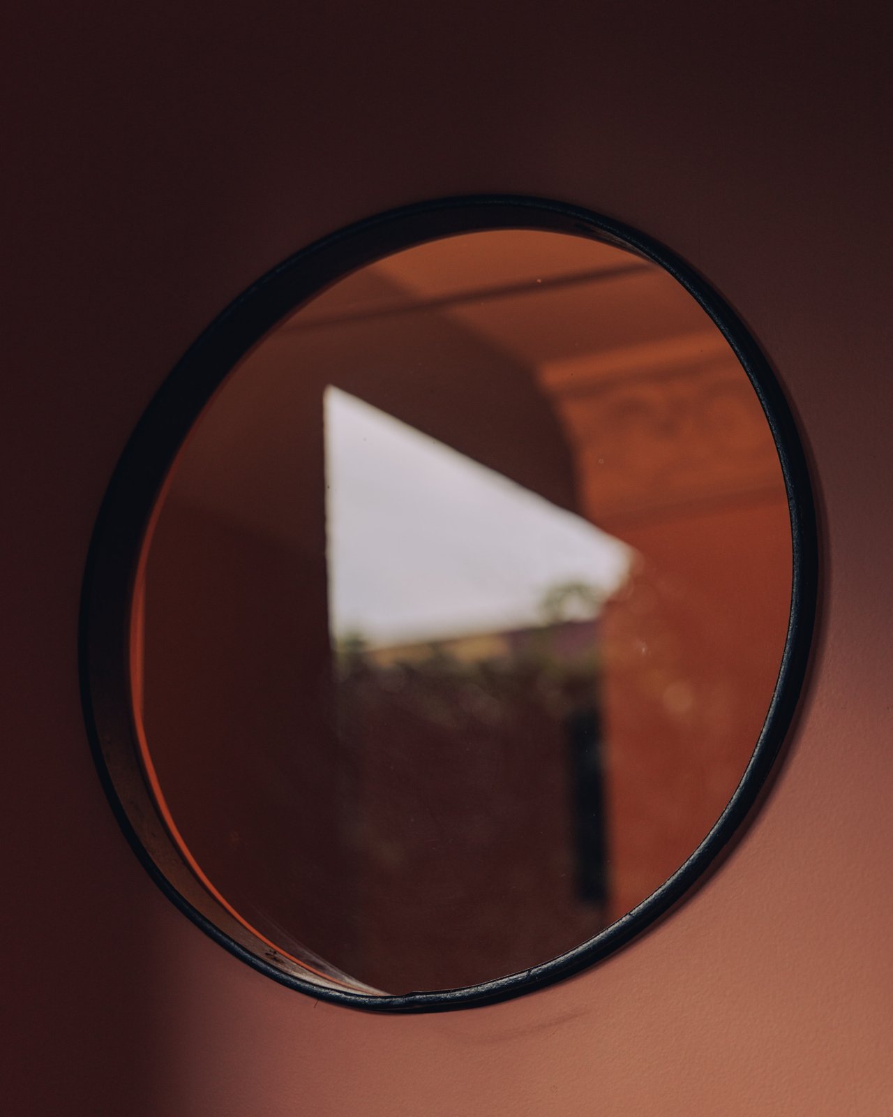 A round black-framed mirror reflecting the terracotta room, Ignacia Guest House