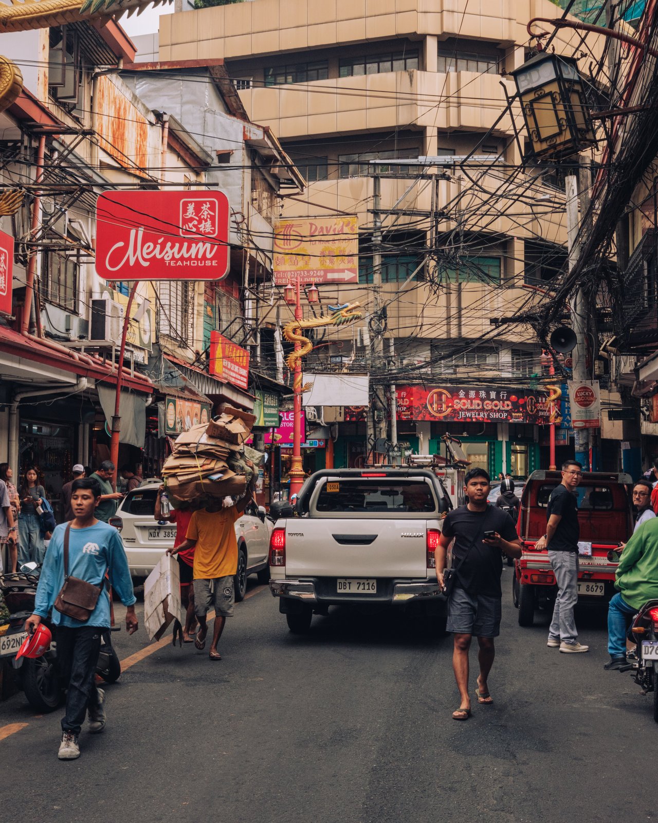 A crowded Binondo street with a 'Meisum' teahouse sign, a porter carrying a sack through traffic, Manila Chinatown