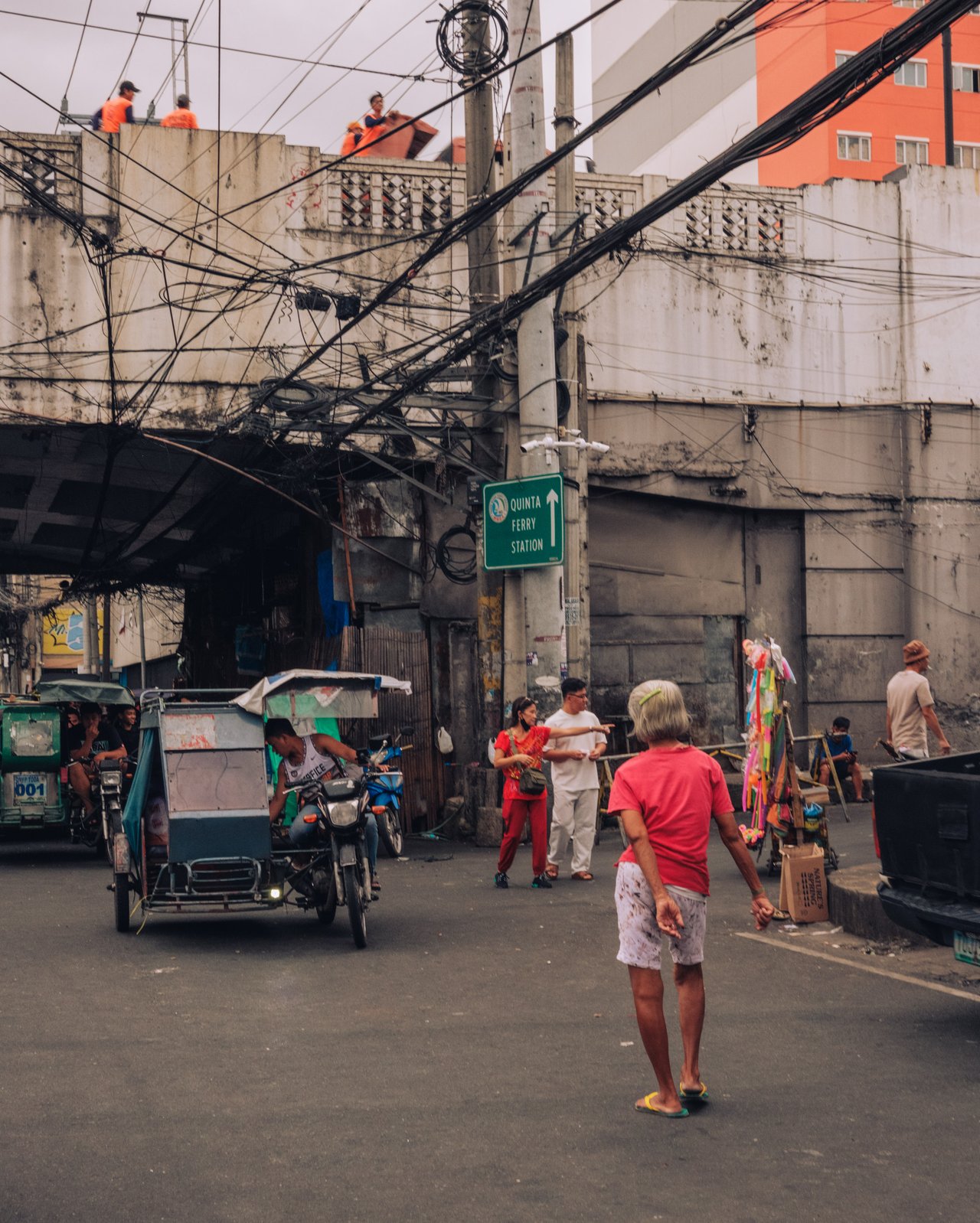 A tricycle and children on a wire-strung street, workers on scaffolding above, Manila