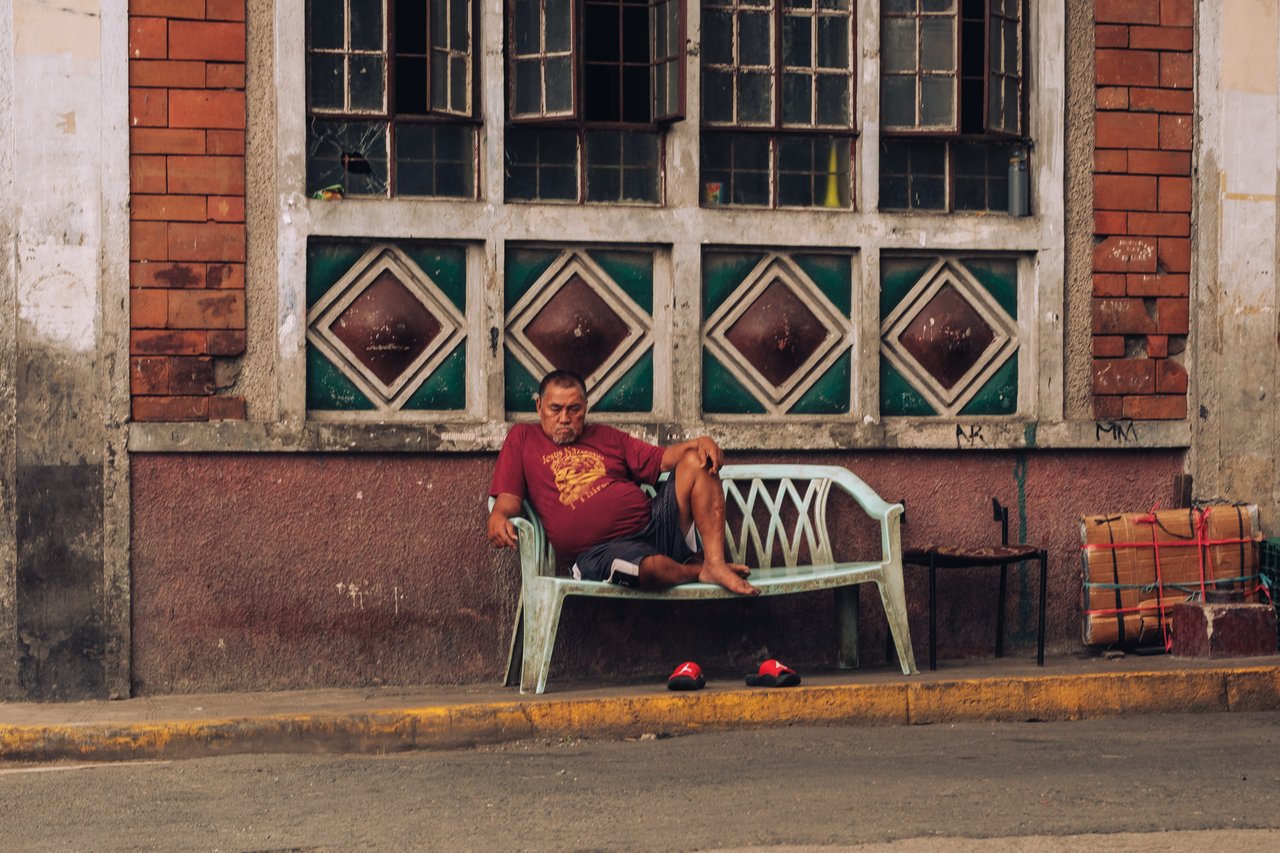 A man in a Superman shirt resting on a plastic bench against an old tiled facade, Manila