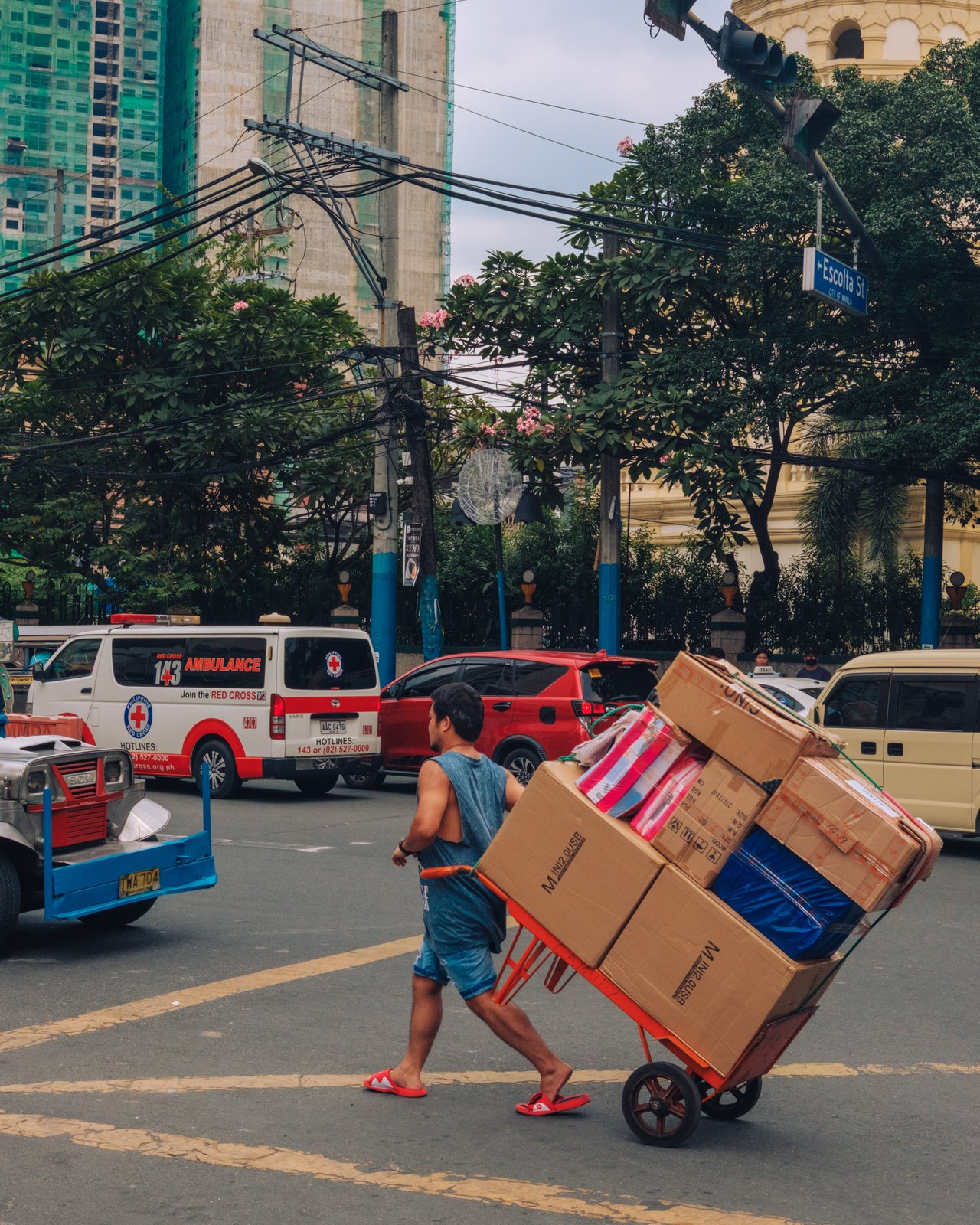 A porter pushing a hand truck stacked with boxes across the street, an ambulance behind, Manila