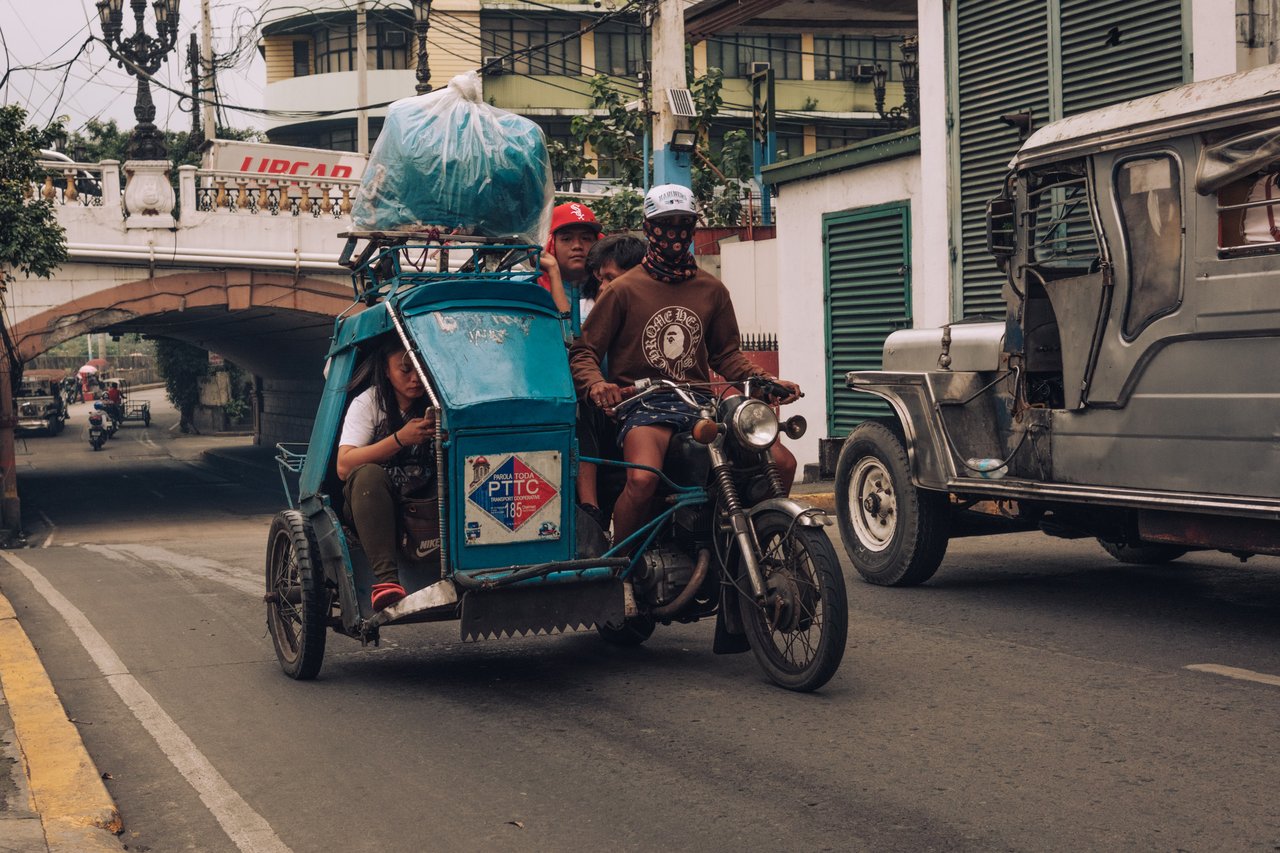 A loaded tricycle passing under a stone bridge as a jeepney rolls by, Manila