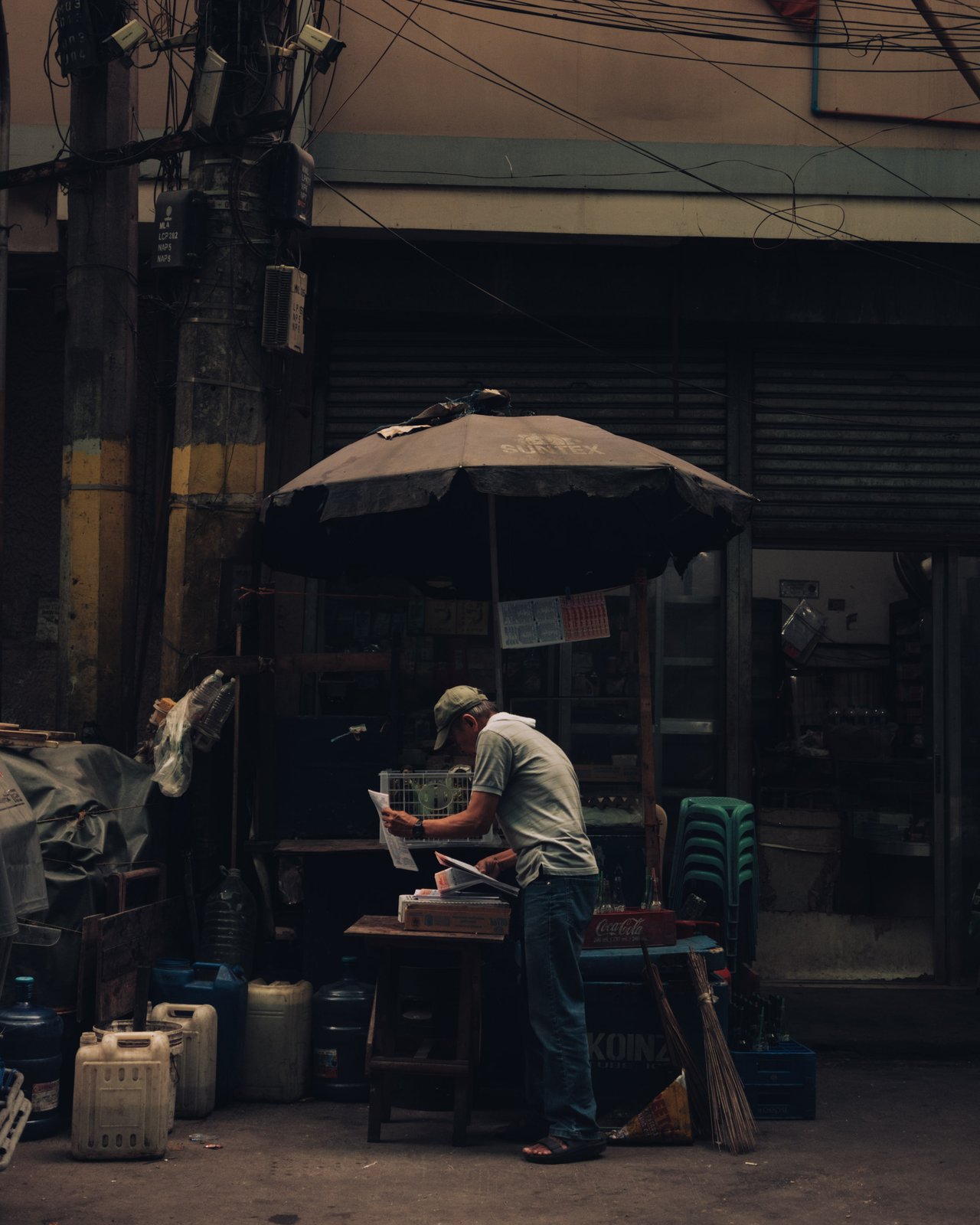 A vendor reading at his sidewalk stall under a black umbrella among shuttered shops, Manila