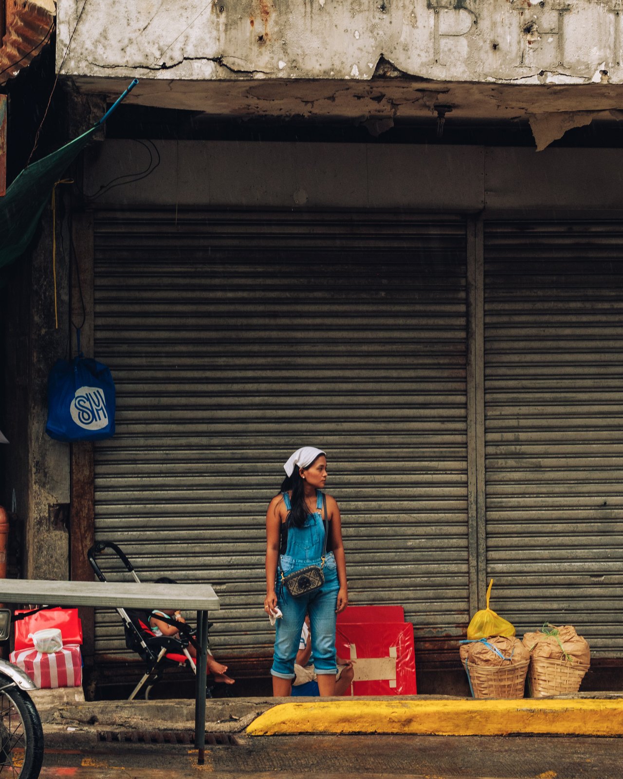 A woman by a roadside stall in front of a rusted shutter, baskets at her feet, Manila