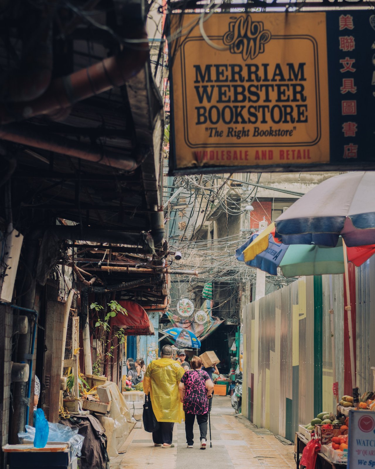 A narrow market alley beneath a 'Merriam Webster Bookstore' sign, shoppers under umbrellas, Manila