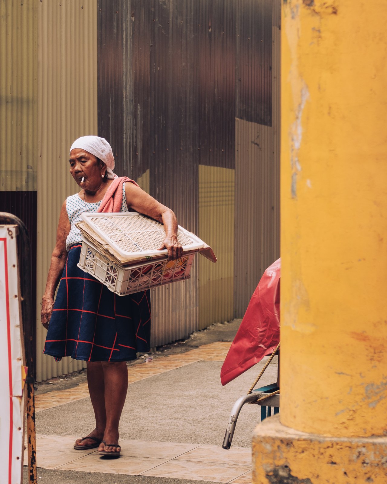 An older woman carrying a plastic crate on her hip past a yellow column, Manila