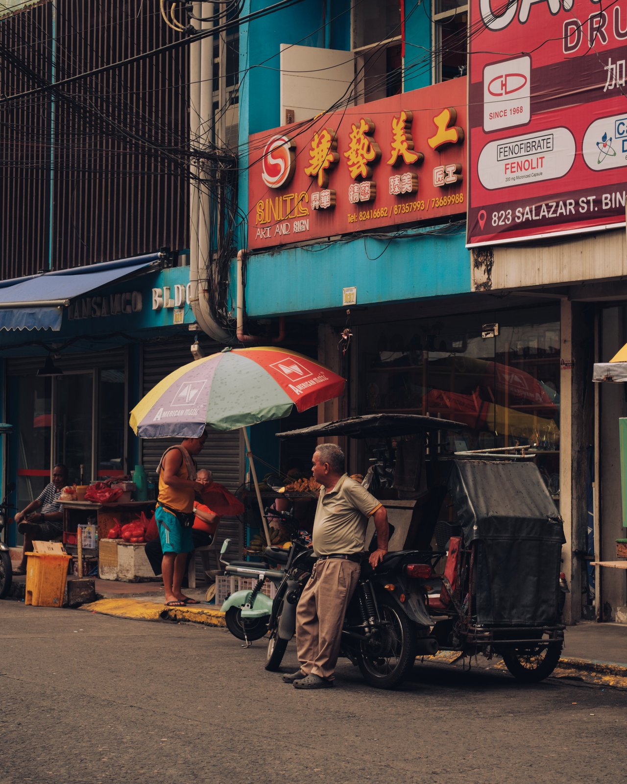 A man beside his parked tricycle at a Binondo corner, Chinese signage and a fruit stall behind, Manila