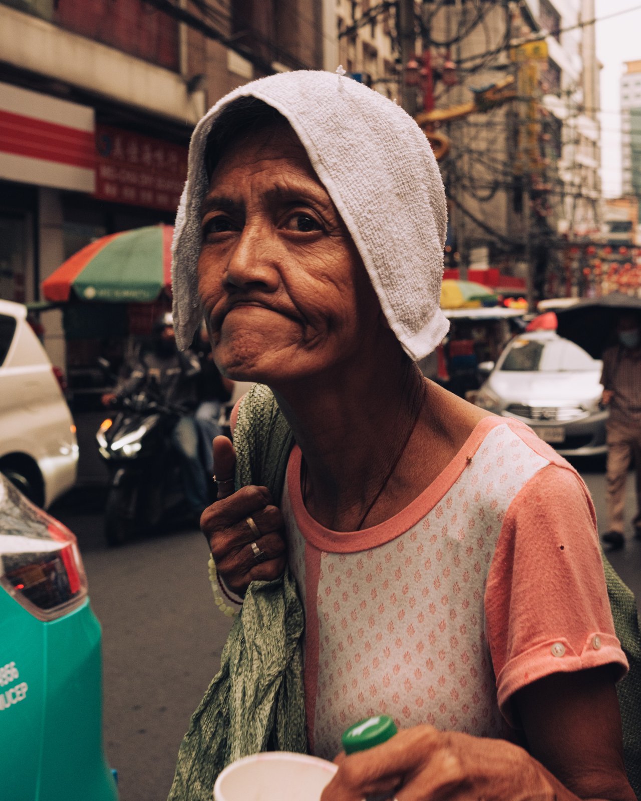 A portrait of an older woman with a towel over her head holding a cup in a busy Chinatown street, Manila