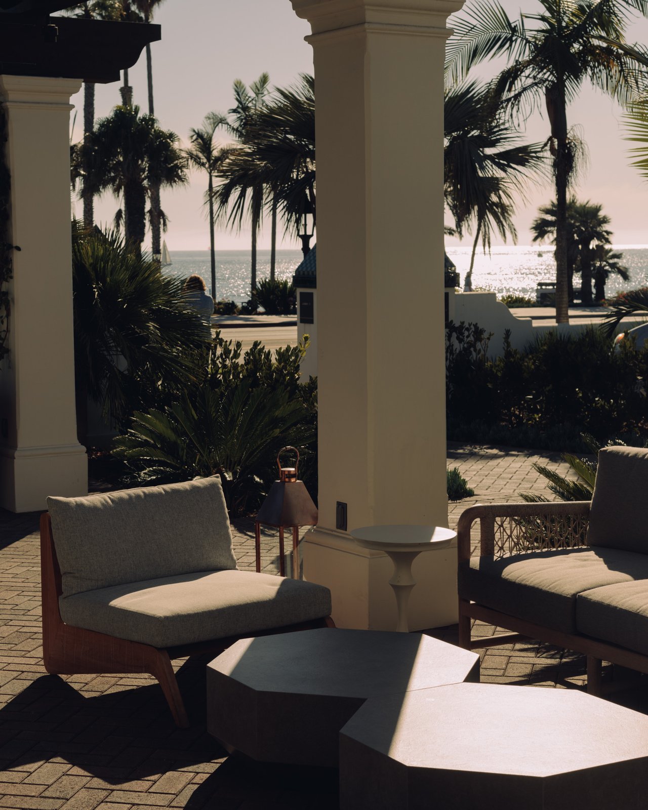 A shaded terrace lounge under the arcade, low seating framed by palms with the ocean beyond