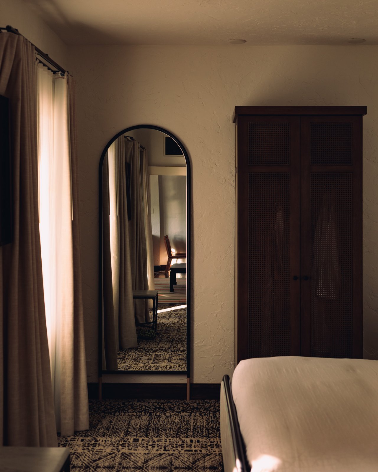 A full-length arched mirror and a dark wood armoire in a light-filled guest room