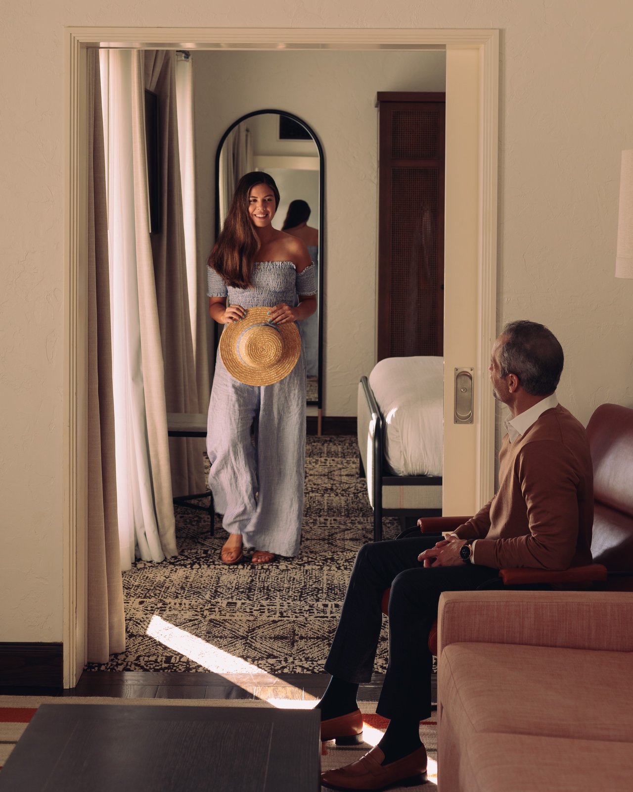 A woman with a straw hat framed in a doorway mirror while a man waits on the sofa of a suite