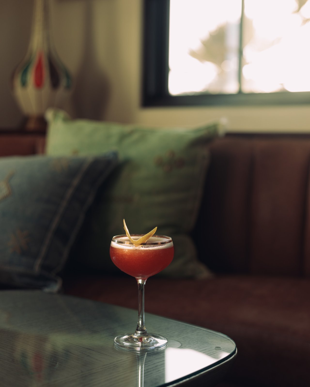 A cocktail with an orange twist on a lounge table, green cushions behind