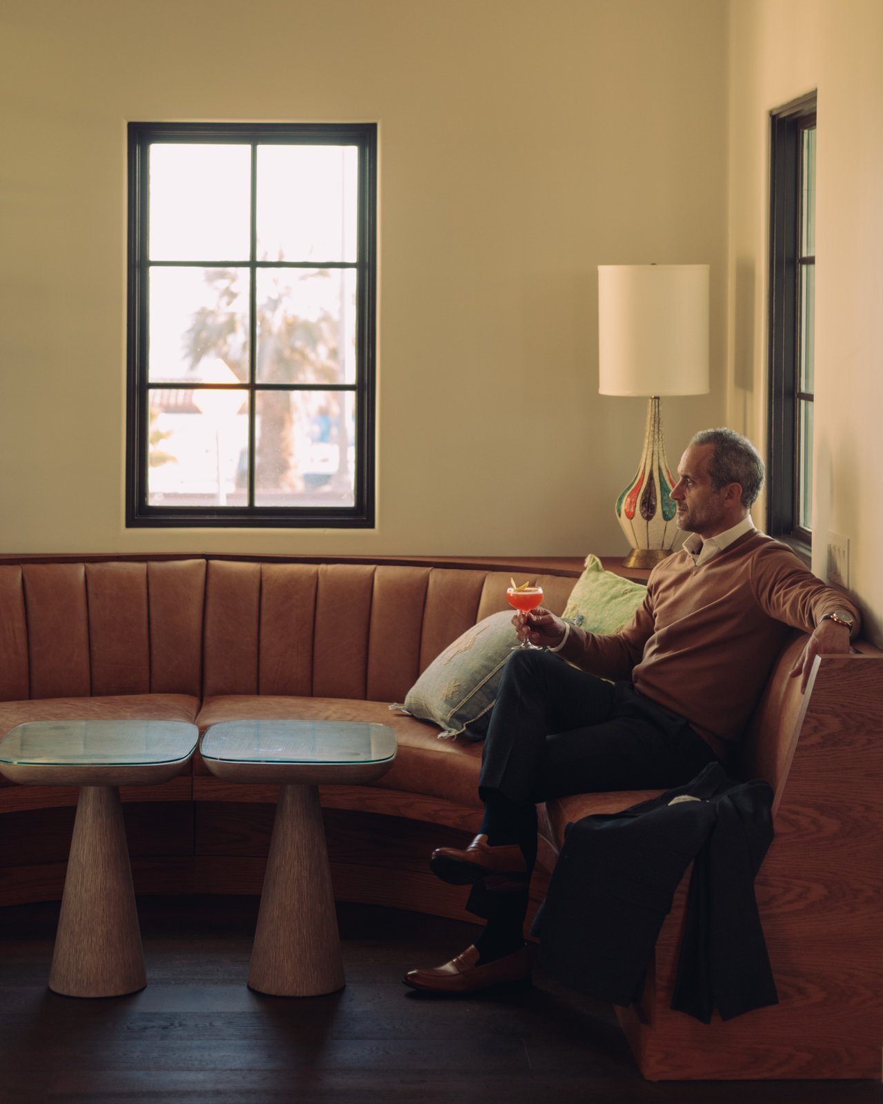 A man with a cocktail on a curved banquette by a bright window in the hotel lounge