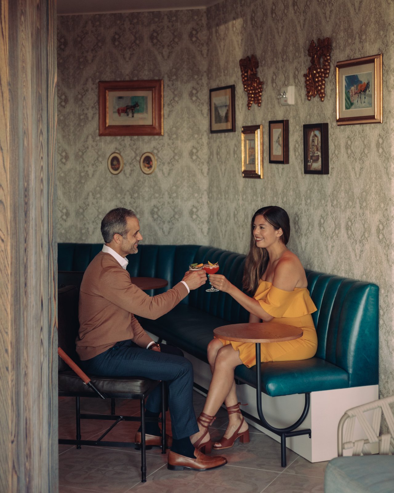 A couple toasting cocktails in a green-leather booth beneath a gallery wall of framed art