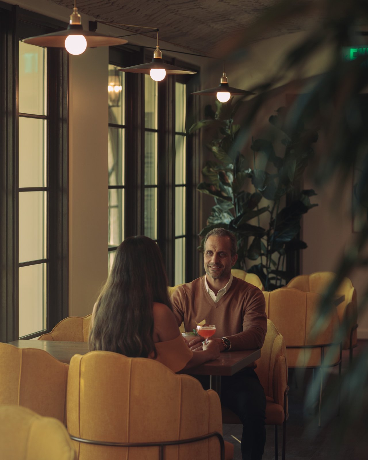 A couple with cocktails in mustard-yellow tub chairs by tall windows at the Mar Monte Hotel