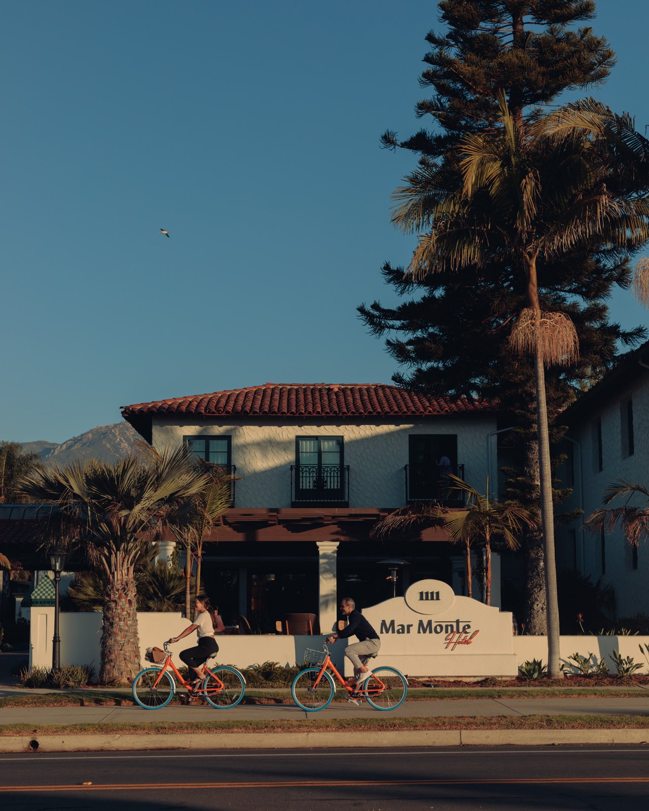 Two cyclists passing the Mar Monte Hotel on Cabrillo Boulevard, palms above the red-tile roofline