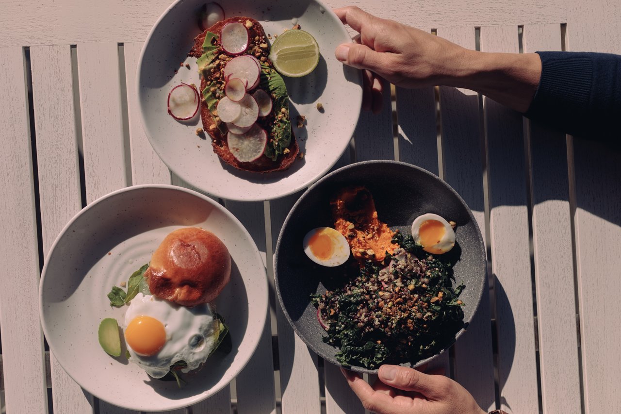 An overhead brunch spread — avocado toast, a burger with egg and a grain bowl — hands reaching in