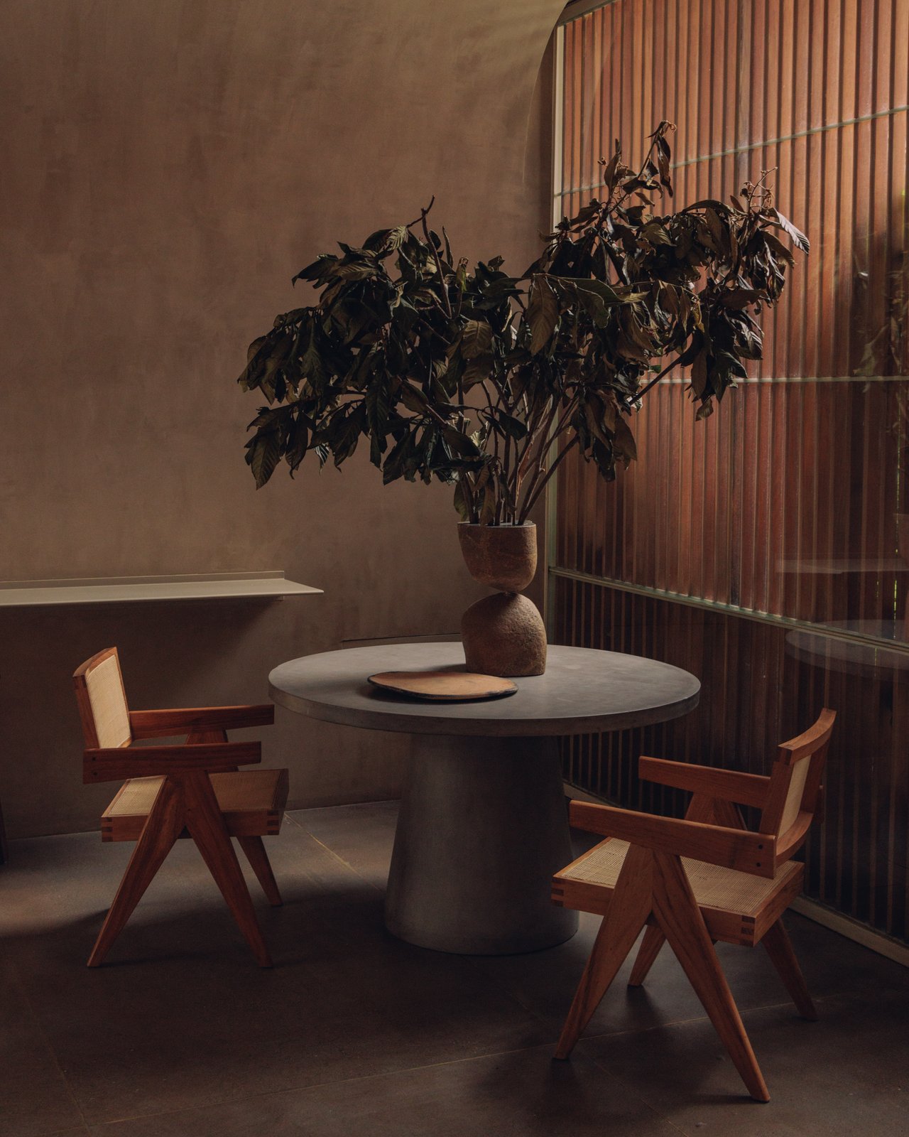 A caned Chandigarh-style chair at a round table by a fluted-wood screen, late sun across the floor