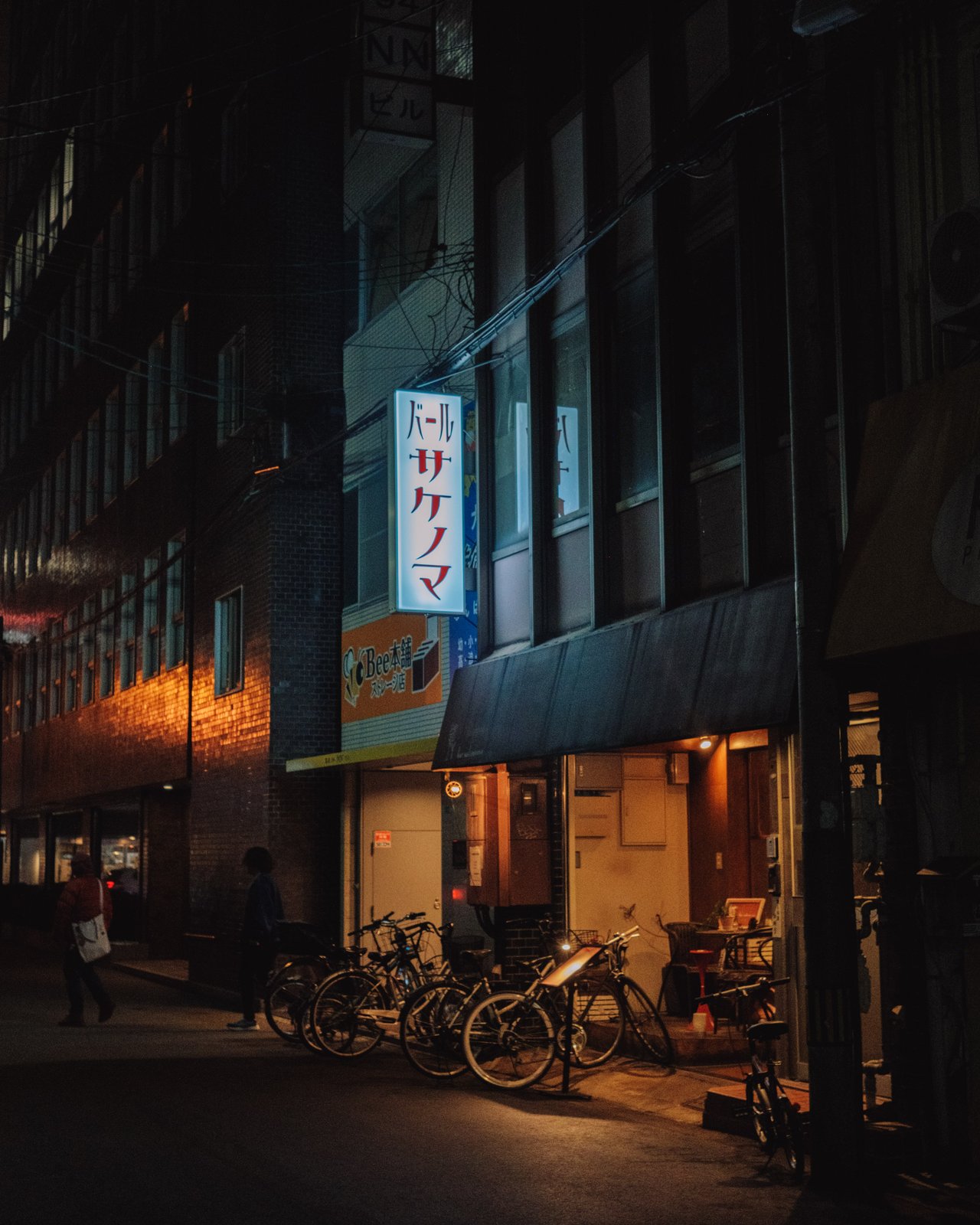 A night street with an izakaya and a sake bar's neon sign, bicycles parked outside, Osaka