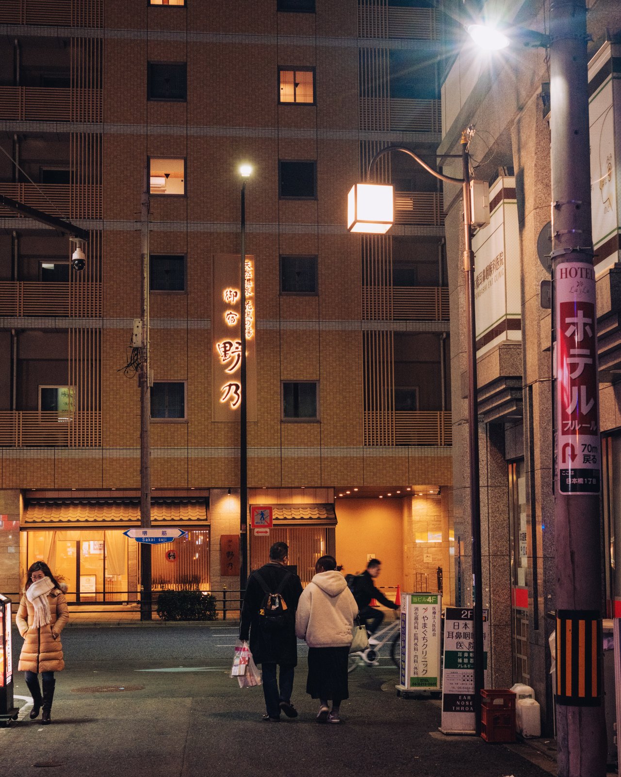 An evening street corner with people walking past an apartment block and a hotel sign, Osaka
