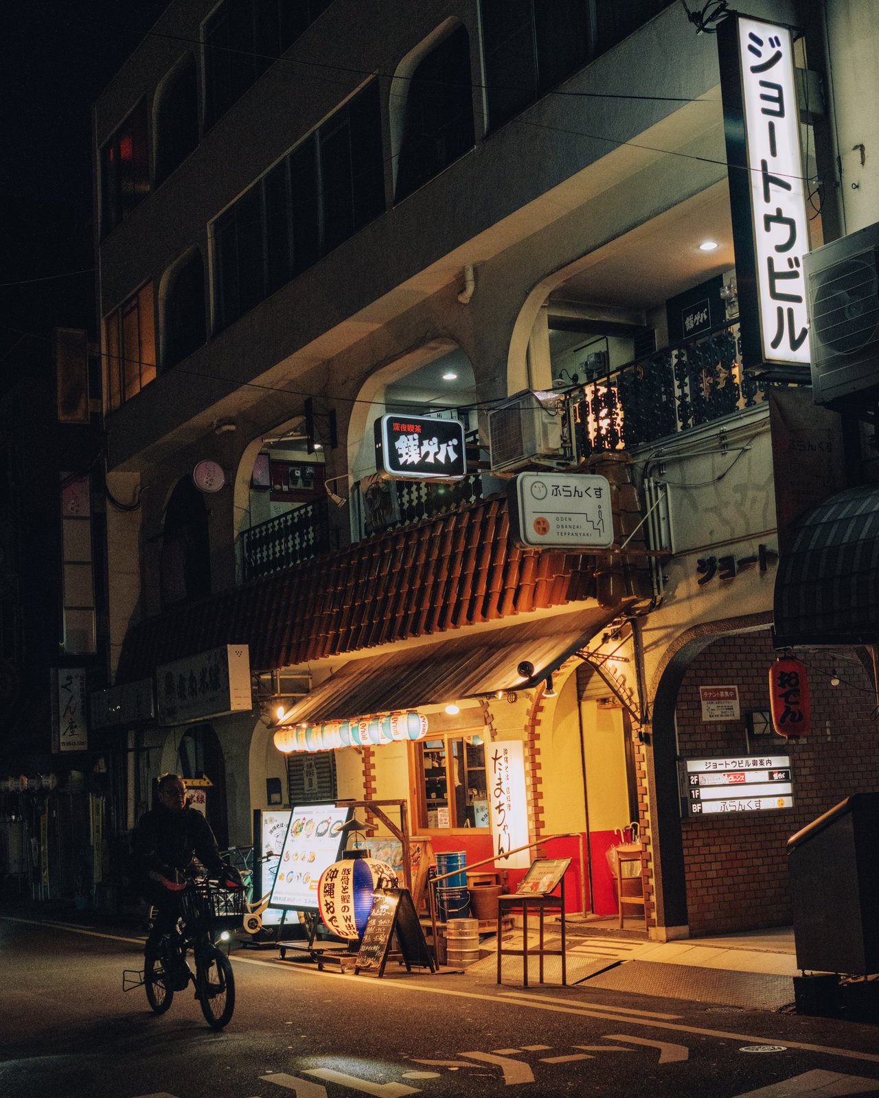 A man cycling down a narrow night street lined with izakayas, Osaka