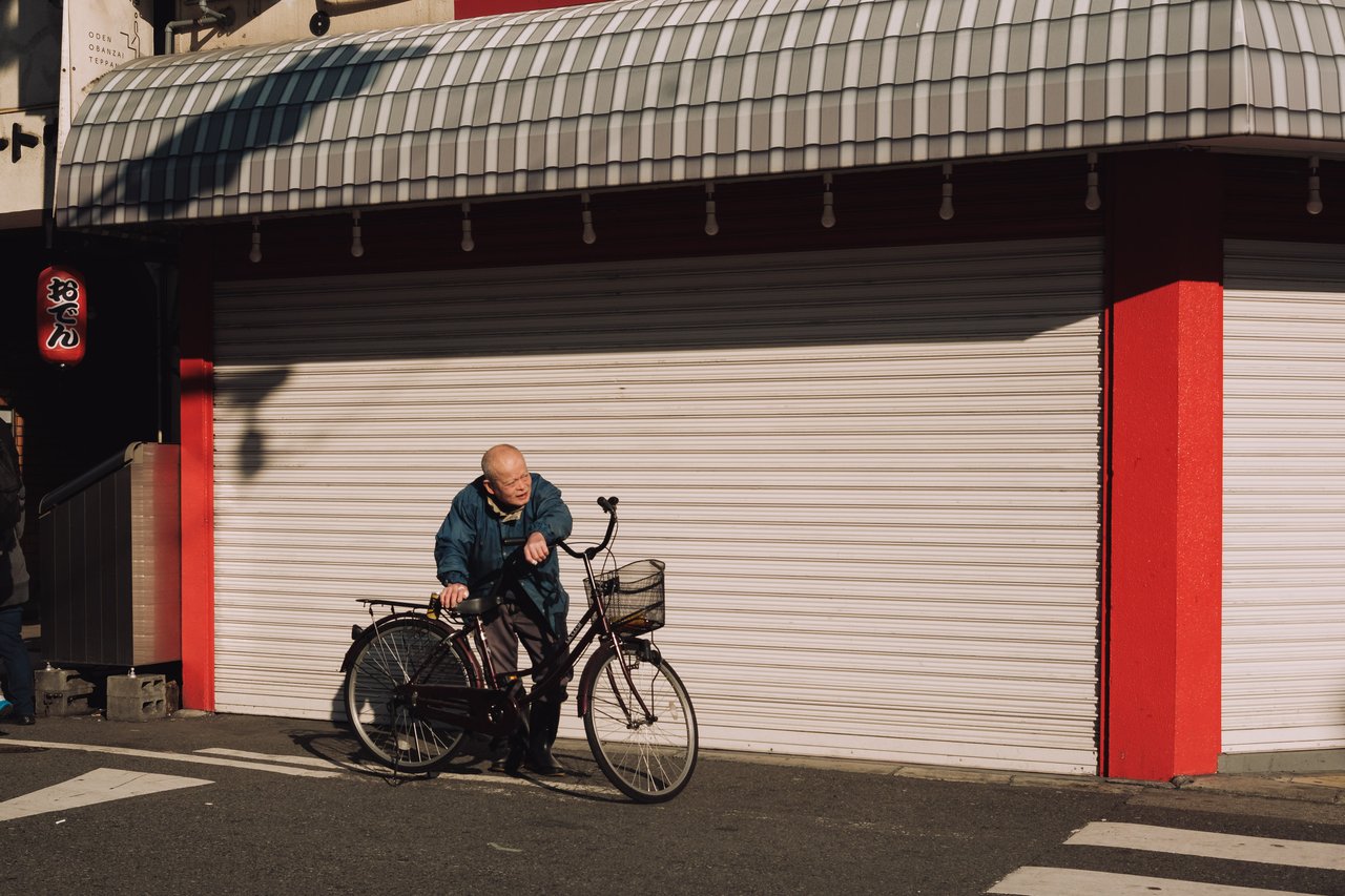 An older man walking his bicycle past a closed red-and-white shutter, Osaka