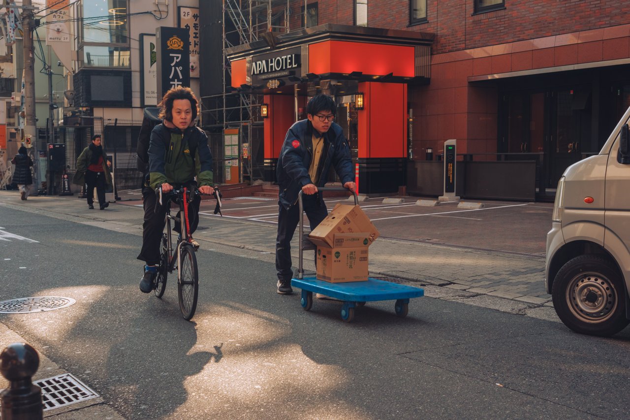 A cyclist and a man pushing a small wood cart crossing a street by an APA Hotel, Osaka