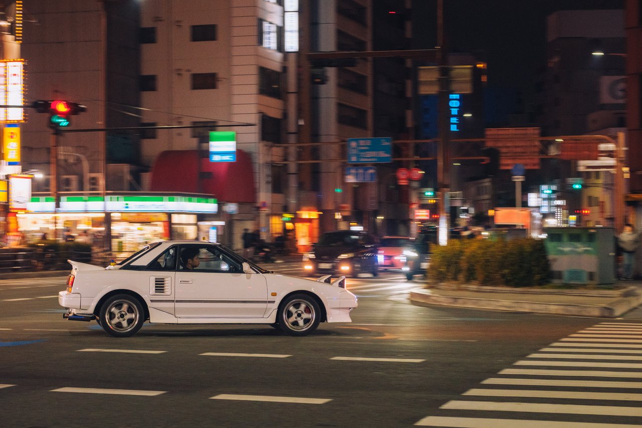 A white classic sports car crossing a neon night intersection, Osaka