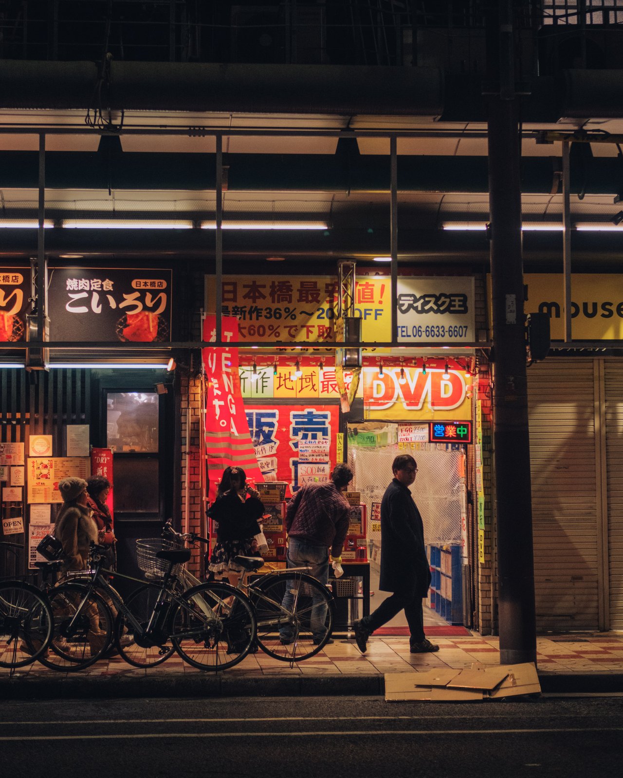 A night shopfront of red lanterns and a DVD shop, bicycles and passers-by, Osaka